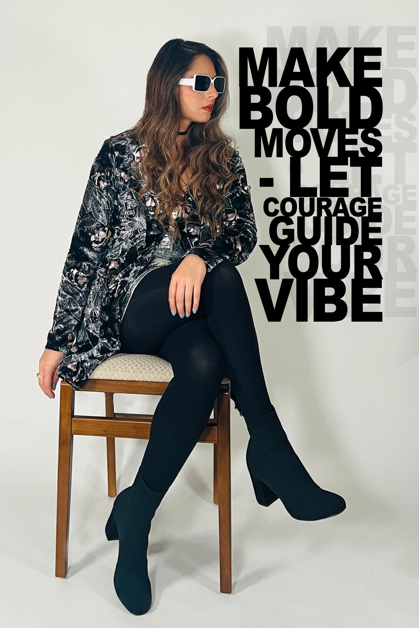 Woman sitting on a chair wearing a black and silver patterned top, black leggings, and black boots. Text on the wall reads 'MAKE BOLD MOVES - LET COURAGE GUIDE YOUR VIBE'.