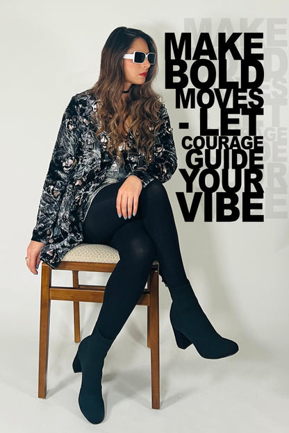 Woman sitting on a chair wearing a black and silver patterned top, black leggings, and black boots. Text on the wall reads 'MAKE BOLD MOVES - LET COURAGE GUIDE YOUR VIBE'.