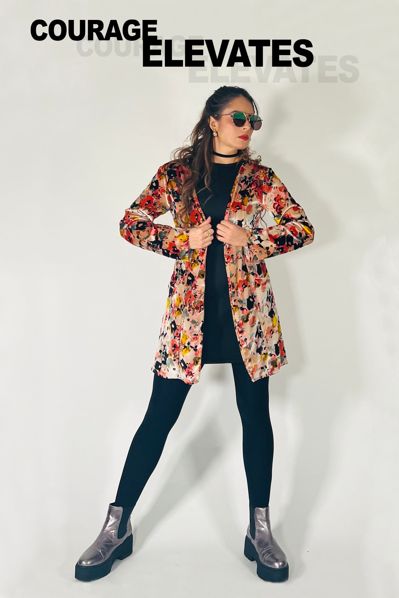 Woman wearing a floral jacket over a black dress with sunglasses, standing against a white background.