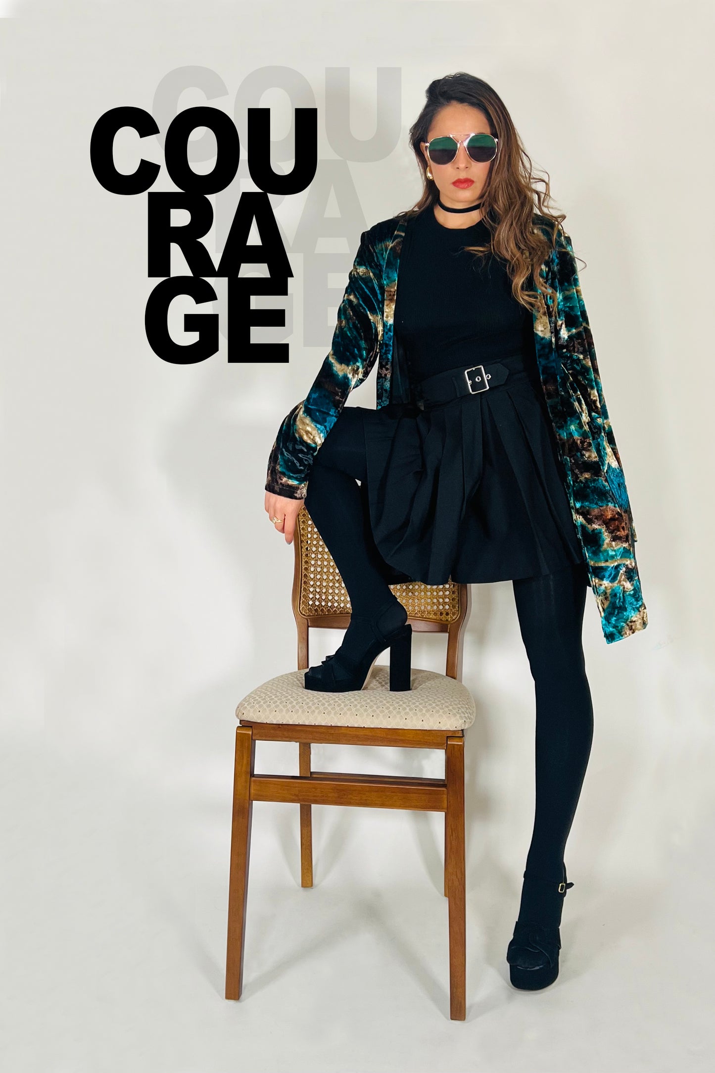 Woman sitting on a chair wearing a floral jacket, black dress, and sunglasses with the word 'COURAGE' displayed.