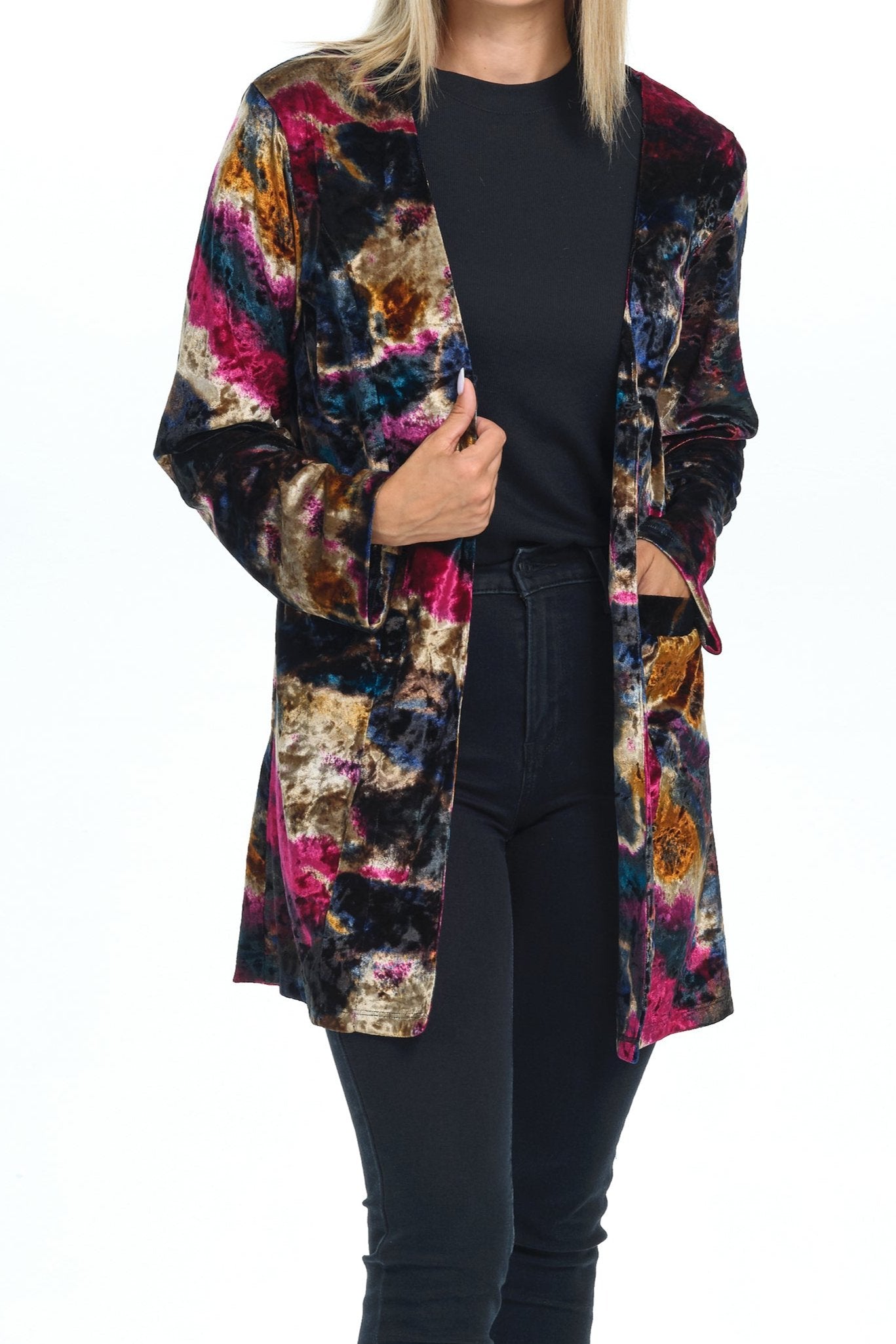 Front view full body of blond-blue-eyes-mid-20s model standing on white background and looking to the camera with one hand on pocket wearing a velvet cardigan with long sleeves and two pockets with black gold pink tiedye printing and all black pants-blouse-sandals outfit