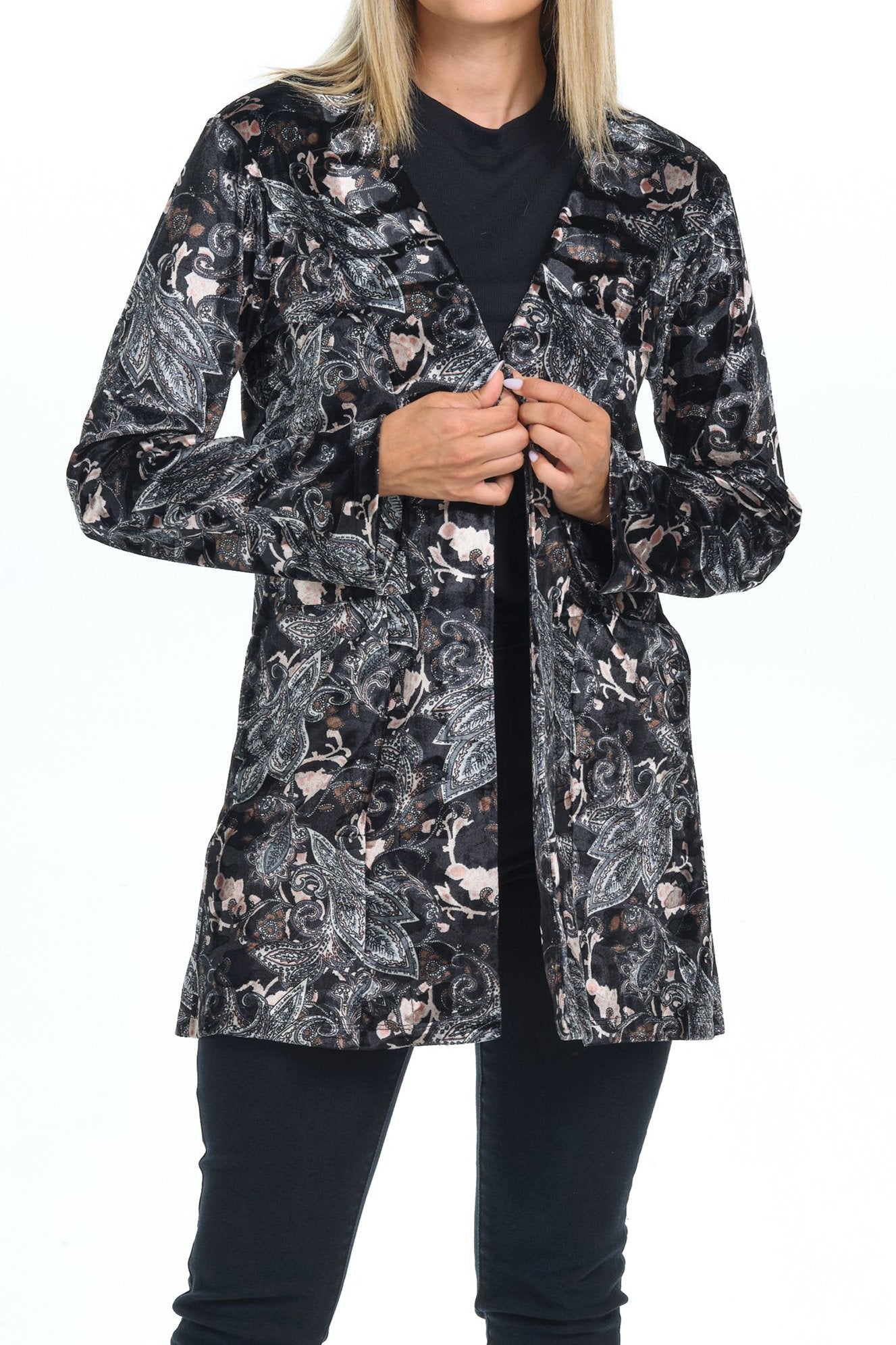 Front view full body of blond-blue-eyes-mid-20s model standing on white background and  looking to the left and holding the sides of the cardigan wearing a velvet cardigan with long sleeves and two pockets with black grey silver paisley printing and all black pants-blouse-sandals outfit