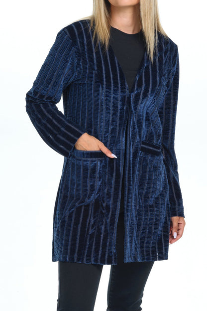 Front view full body of blond-blue-eyes-mid-20s model standing on white background and looking to the left one hand on pocket of velvet cardigan she is wearing with long sleeves and two pockets colored navy blue stripped and all black-pants-blouse-sandals outfit