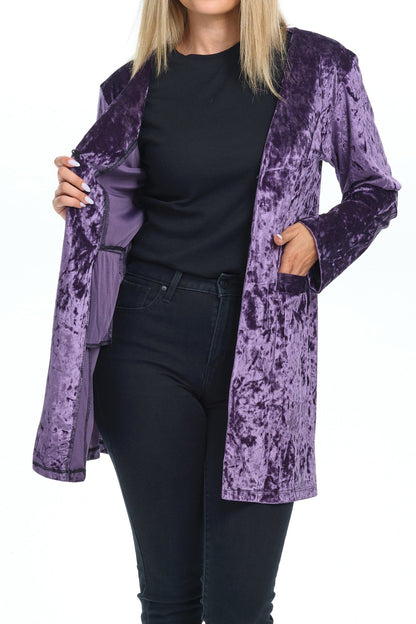 Front view full body of blond-blue-eyes-mid-20s model standing on white background and looking to the camera crossed-legs both hands on the pockets of the velvet cardigan she is wearing with long sleeves and two pockets colored purple shine eggplant and all black pants-blouse-sandals outfit