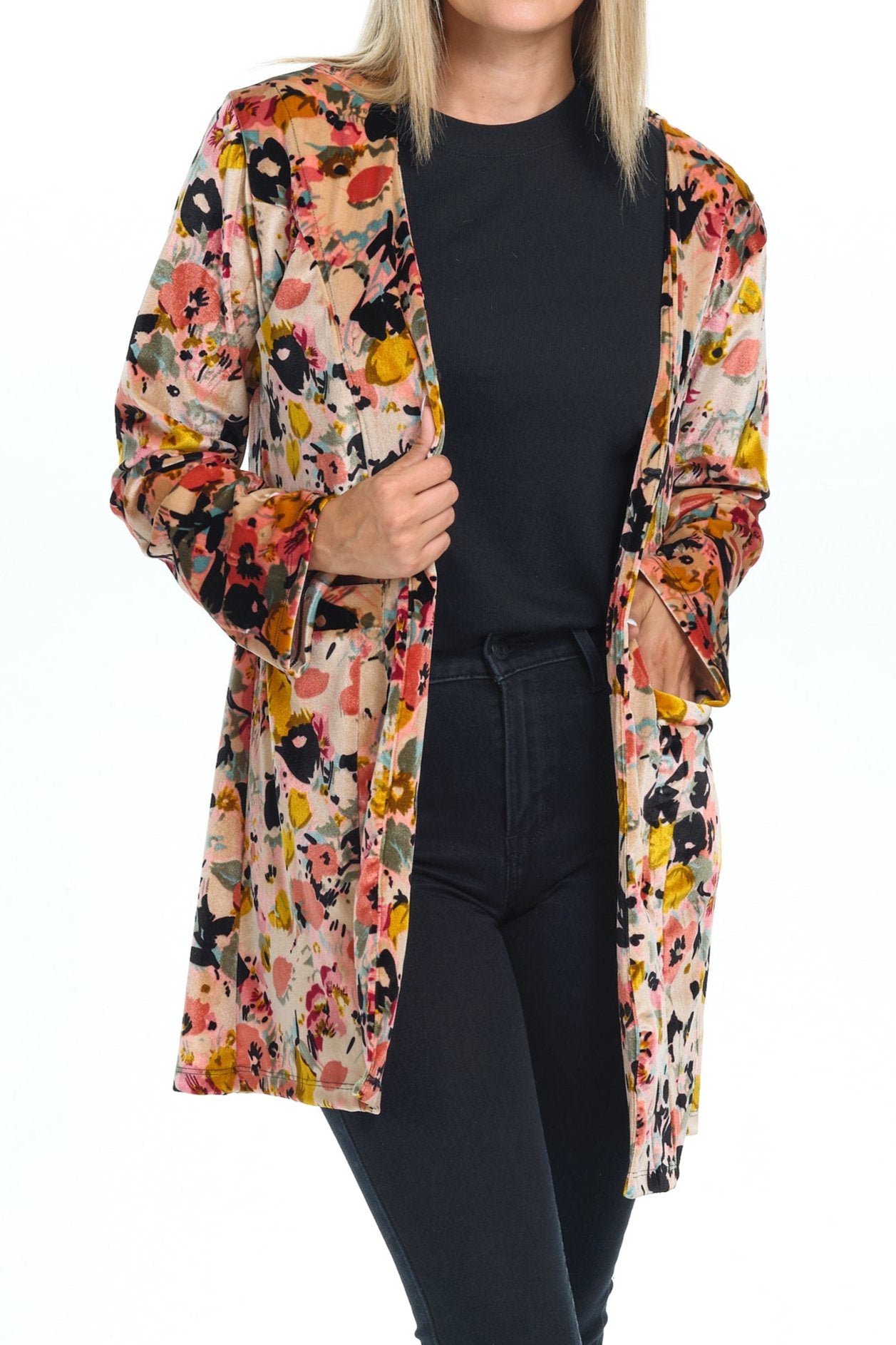 Front view full body of blond-blue-eyes-mid-20s model standing on white background and looking to the camera crossed-legs wearing a velvet cardigan with long sleeves and two pockets with rosepink black yellow printing  and all black pants-blouse-sandals outfit